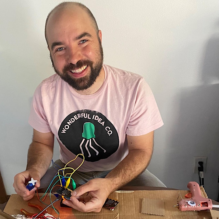 Ryan smiling whilst holding a motor, microbes and wires. There is a glue gun to his side.