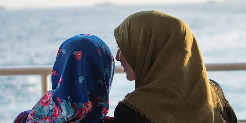 Photo from behind of a woman in a head scarf leaning to speak with another woman wearing a headscarf