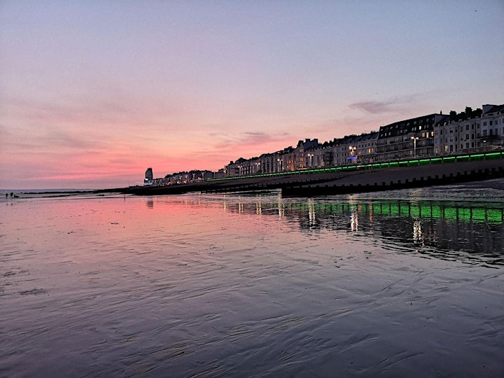 Hastings Seafront at sunset. The pink and purple sky is reflected in a calm sea. The lights in the buildings are illuminated.