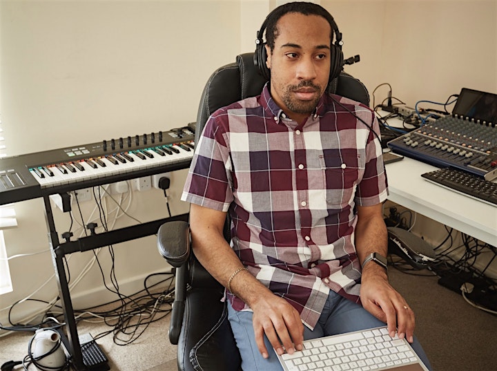 A portrait photograph of a man with short black hair wearing black headphones and a burgundy and white checked shirt sitting down. There are keyboards and music equipment in the background. 