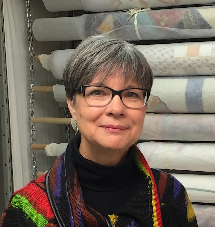 Image of Lucie Heins, a woman wearing glasses with short grey hair. She is standing in front of rolled, stored textiles. 